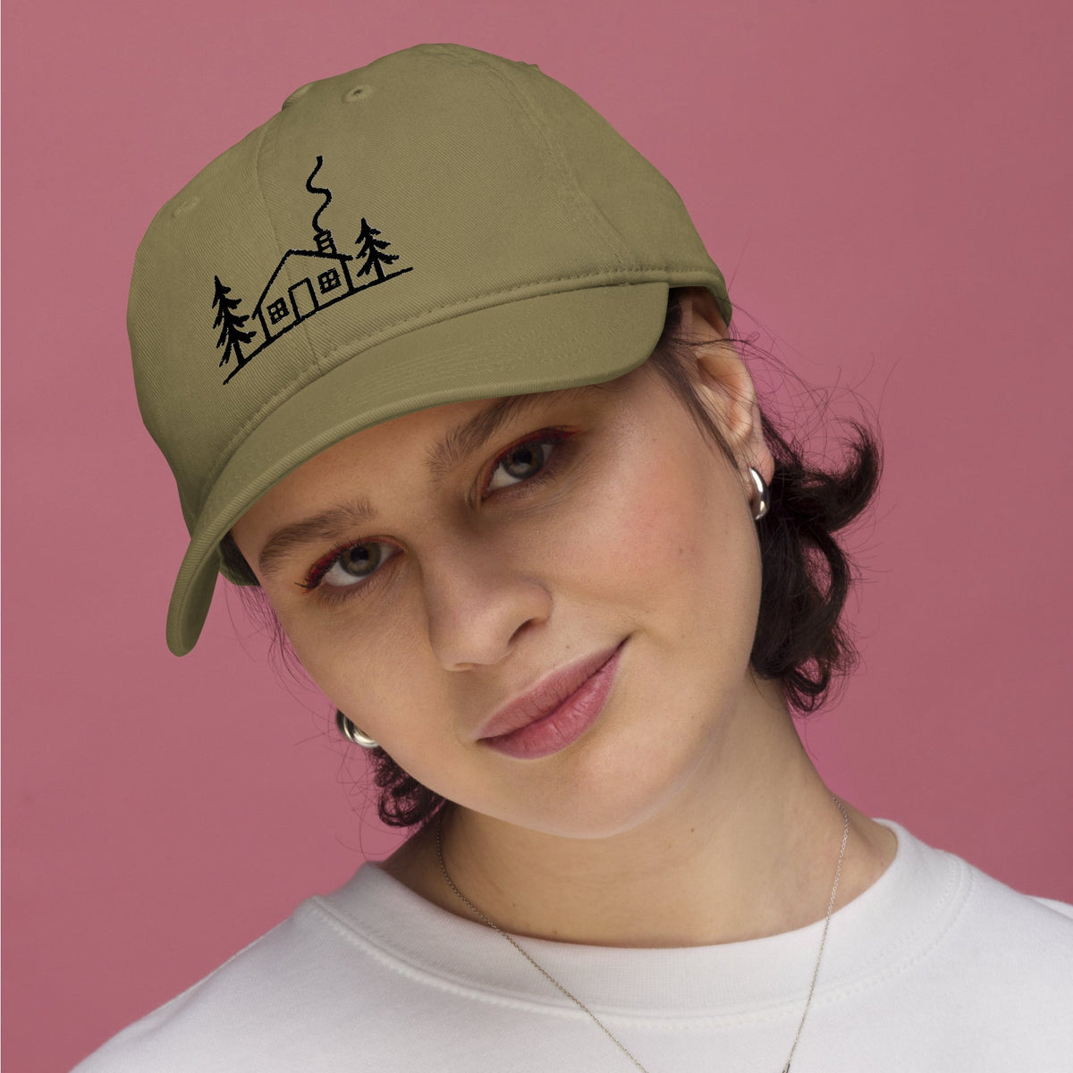 Female model wearing the jungle green Meet Me at the Cabin baseball hat by Klemm & Co., featuring the black embroidered cabin and pine tree design.