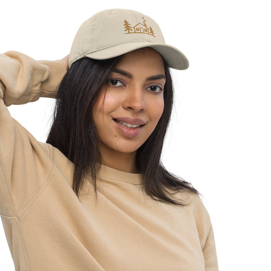 Model wearing the oyster Meet Me at the Cabin baseball hat by Klemm & Co., featuring gold embroidered cabin and pine tree artwork.