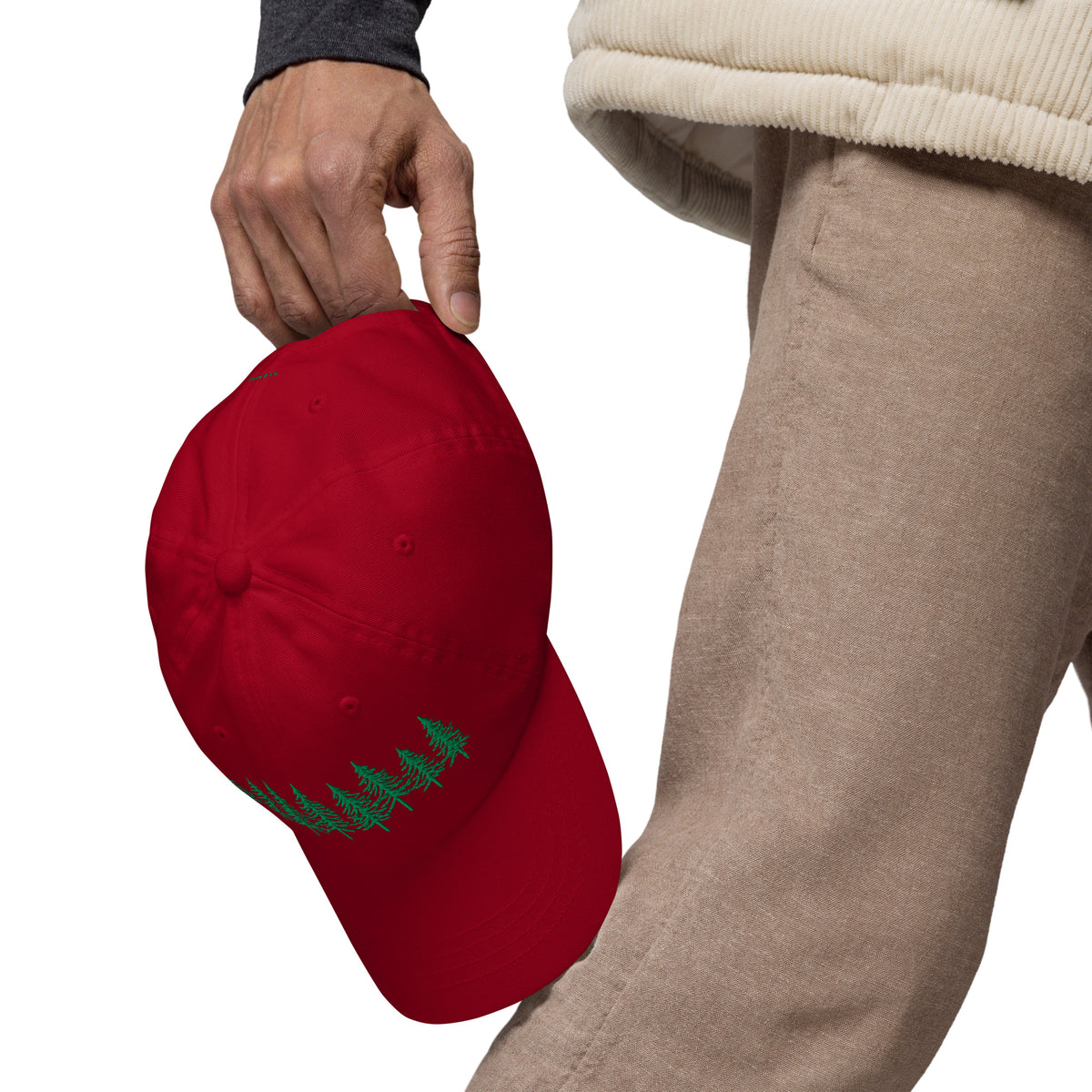 Male model holding the cranberry Into the Pines baseball hat by Klemm & Co. at his side, showing the embroidered pine tree design in green thread.