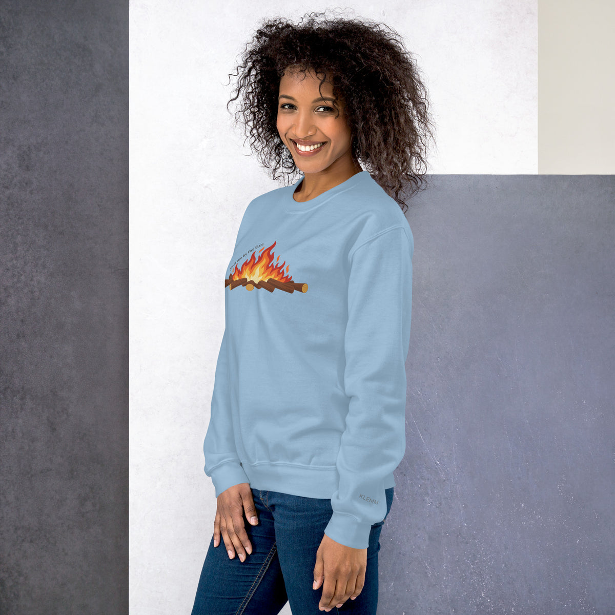 Female model wearing the light blue Meet Me by the Fire crewneck sweatshirt by Klemm & Co., front-left view showing the printed campfire design and KLEMM sleeve branding.