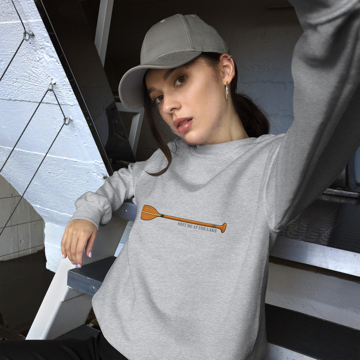 Female model wearing the sport grey Meet Me at the Lake crewneck sweatshirt by Klemm & Co., front view with her left arm raised, showing the printed paddle design.