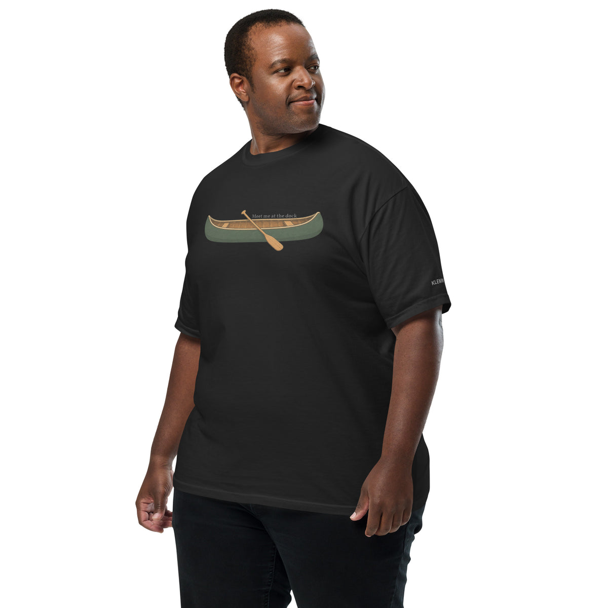 Plus-size male model wearing the black Meet Me at the Dock graphic T-shirt by Klemm & Co., front-left view showing the printed canoe design and KLEMM sleeve branding.