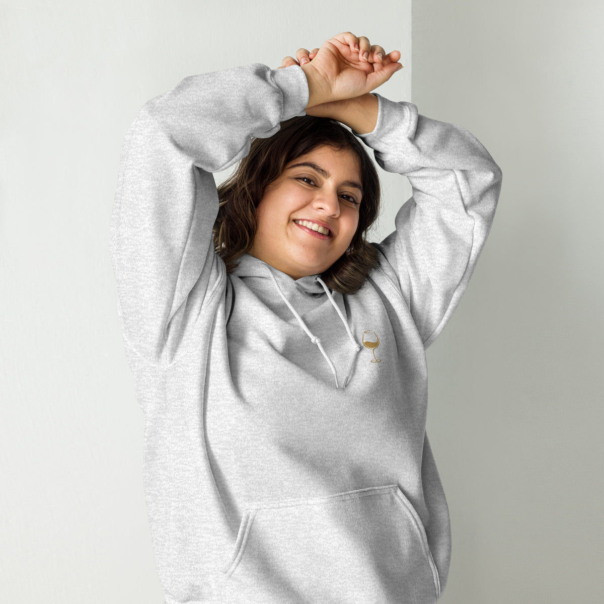 Female model wearing the ash Poured for This wine hoodie by Klemm & Co., front view with arms raised, showing the gold embroidered wine glass design.