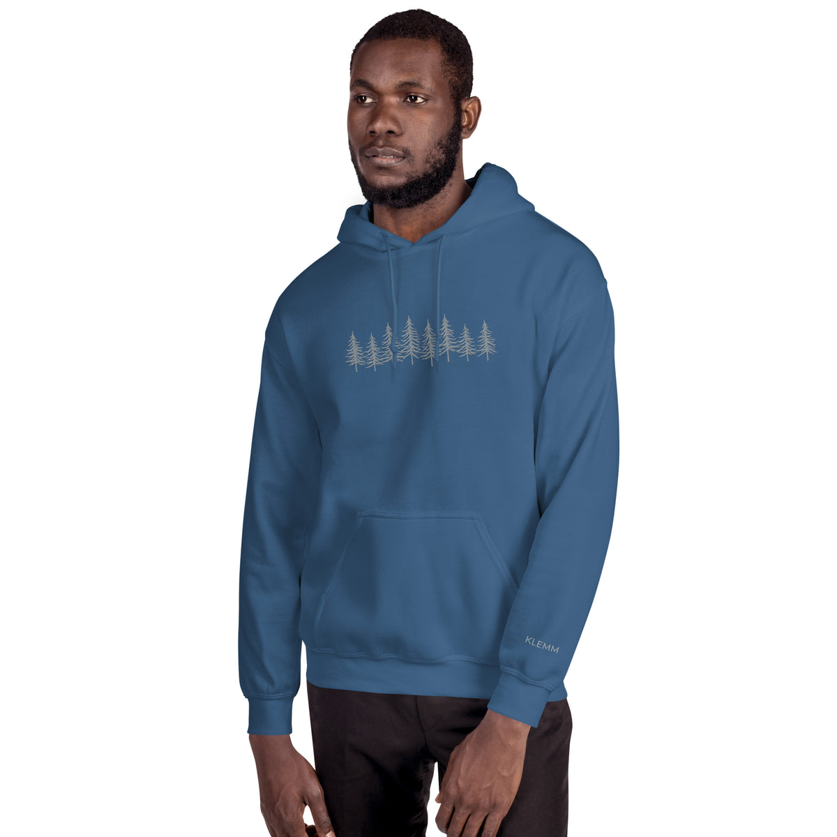 Male model wearing the indigo blue Into the Pines embroidered hoodie by Klemm & Co., front-left view showing the grey embroidered pine tree design and KLEMM sleeve branding.