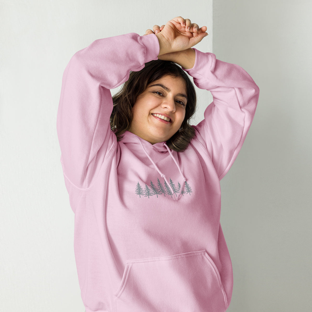 Female model wearing the light pink Into the Pines embroidered hoodie by Klemm & Co., front view with arms raised, showing the pine tree design embroidered in grey thread.
