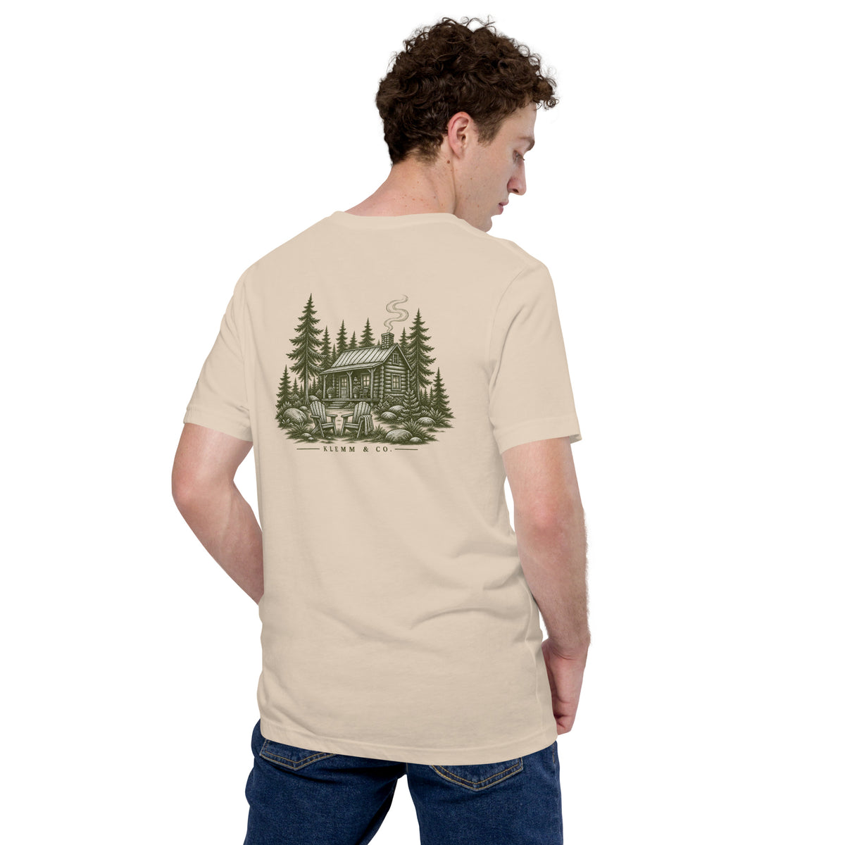 Model wearing the soft cream Meet Me at the Cabin graphic T-shirt by Klemm & Co., showing the woodland cabin back design.