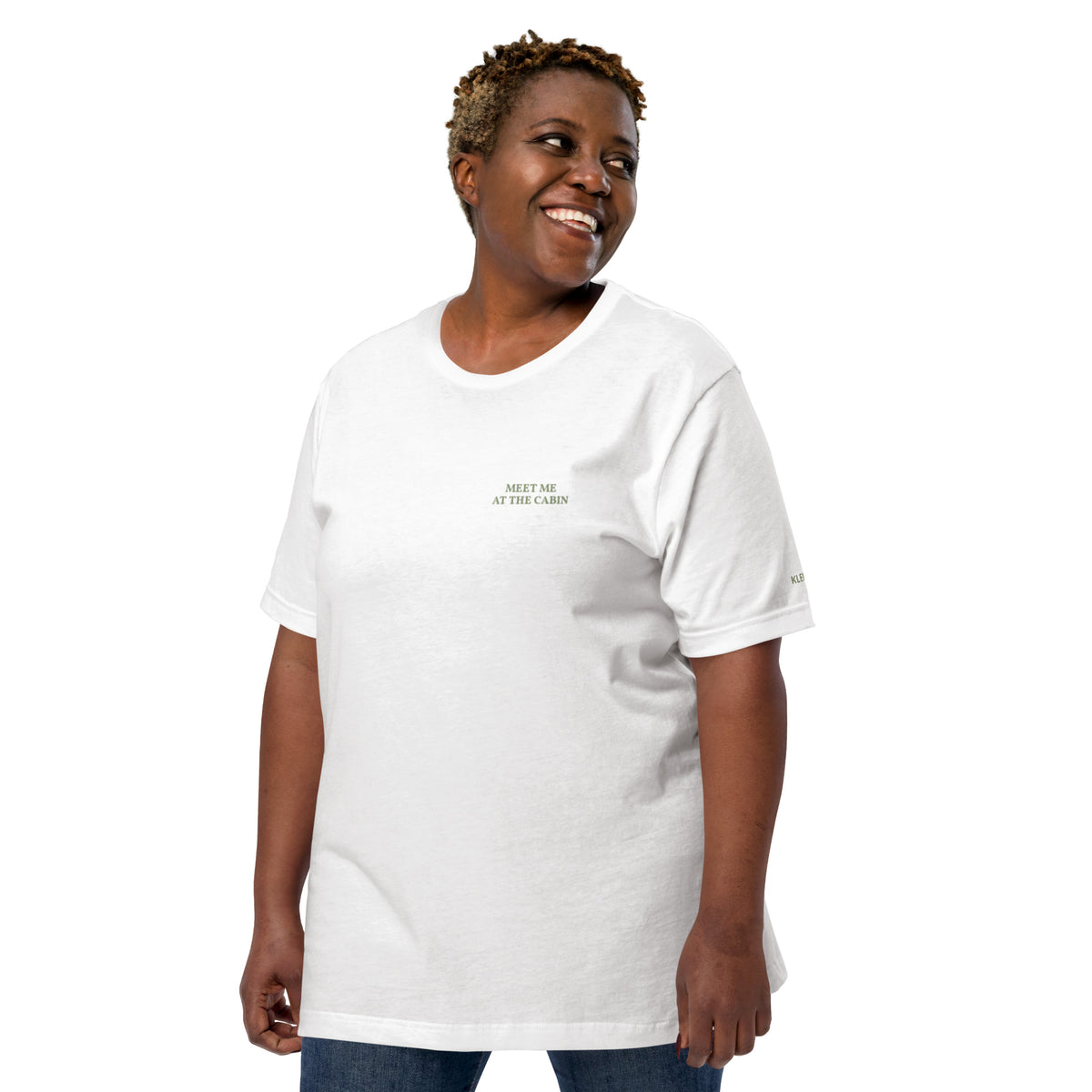 Model wearing the white Meet Me at the Cabin graphic T-shirt by Klemm & Co., front view showing the subtle green chest text.